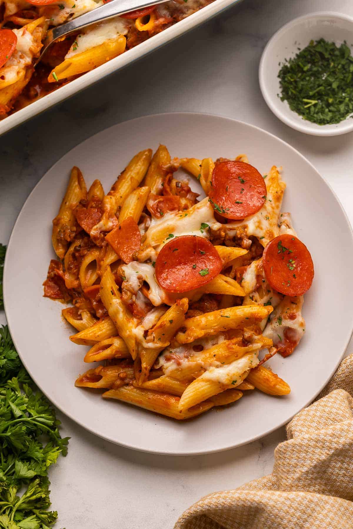 This Pizza Pasta Bake is cheesy, hearty, and loaded with sausage, pepperoni, and marinara. It's like pizza night and pasta night all in one dish! It's make ahead and freezer friendly weeknight meal. #dinner #recipe | make ahead meal | freezer meal | pasta bake | pepperoni pizza | italian sausage | comfort food | easy dinner ideas | dinner recipes a grey plate filled with pizza pasta topped with chopped parsley.