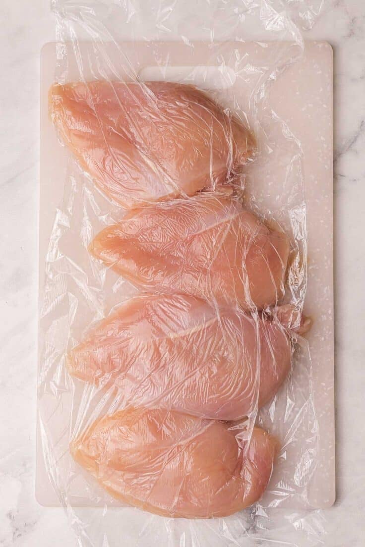 chicken breasts on a cutting board with plastic wrap over.