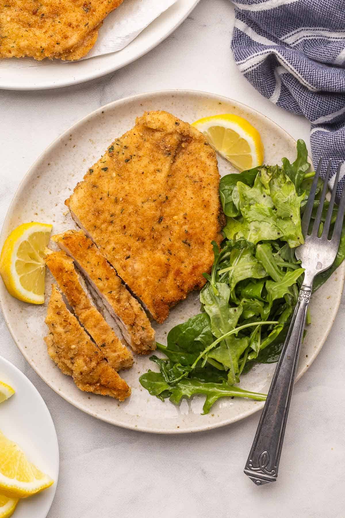 Crispy Chicken Milanese is golden, crunchy, and ready in 30 minutes! Thin chicken cutlets coated in breadcrumbs and pan-fried until perfectly crisp. Perfect with a squeeze of lemon and some fresh greens! #chicken #dinner #recipe | italian food | italian recipes | crispy chicken | breaded chicken | chicken recipes | chicken breast overhead image of chicken milanese on a plate with greens and lemon.