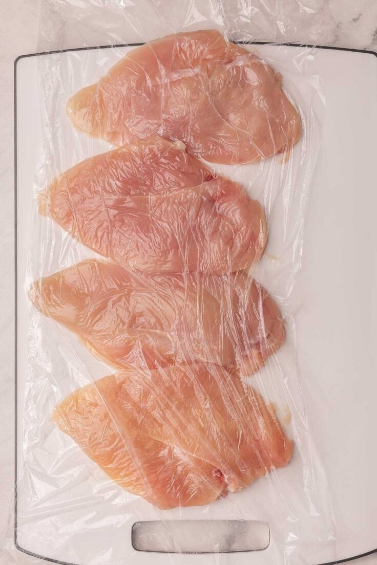 chicken breasts on a cutting board with plastic wrap over top.