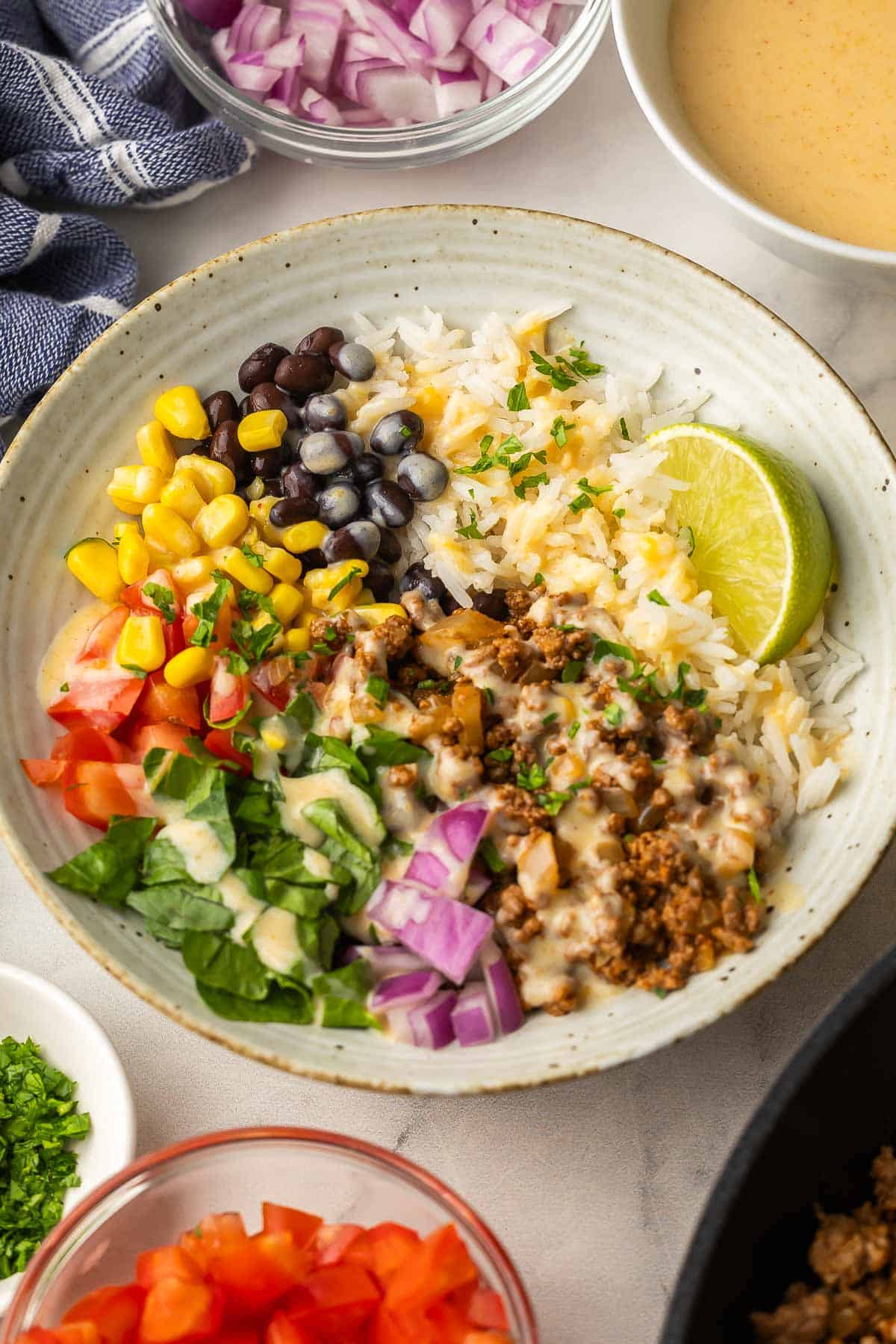 Easy Ground Beef Burrito Bowls with rice, veggies, taco ground beef and creamy nacho cheese sauce! Ready in 35 minutes, totally customizable, and family-approved. #recipe #dinner | easy dinner ideas | ground beef recipes | freezer meals | comfort food | taco night | easy meals | make ahead meals ground beef burrito bowl with corn beans rice tomato red onion and lime wedge.