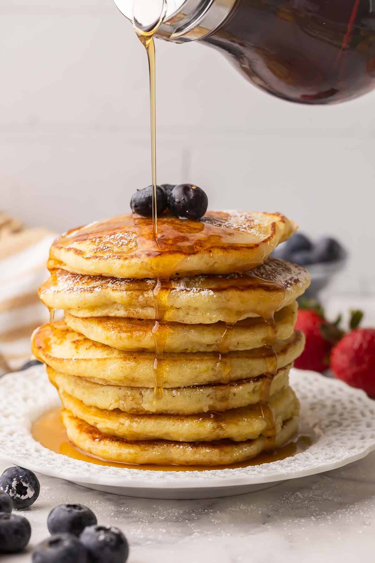 Light, fluffy Lemon Ricotta Pancakes with a burst of citrus and a hint of sweetness! With rich ricotta cheese, lemon juice and lemon zest they are full of flavor and will melt in your mouth. #lemon #pancakes #breakfast #recipe | breakfast recipes | lemon recipes | lemon pancakes | breakfast ideas | brunch recipes | mother's day | easter tall stack of lemon ricotta pancakes with syrup being drizzle over.
