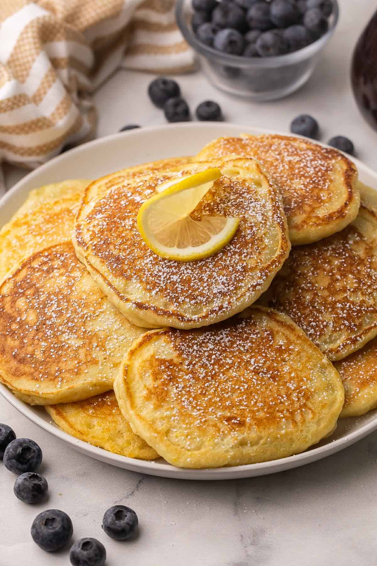 Light, fluffy Lemon Ricotta Pancakes with a burst of citrus and a hint of sweetness! With rich ricotta cheese, lemon juice and lemon zest they are full of flavor and will melt in your mouth. #lemon #pancakes #breakfast #recipe | breakfast recipes | lemon recipes | lemon pancakes | breakfast ideas | brunch recipes | mother's day | easter plate full of lemon ricotta pancakes with powdered sugar and a lime slice on top.