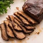 sliced beef brisket on a white cutting board with parsley behind it.