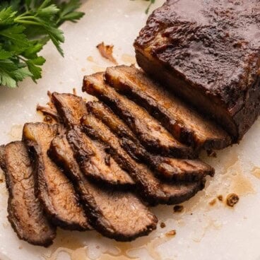 sliced beef brisket on a white cutting board with parsley behind it.