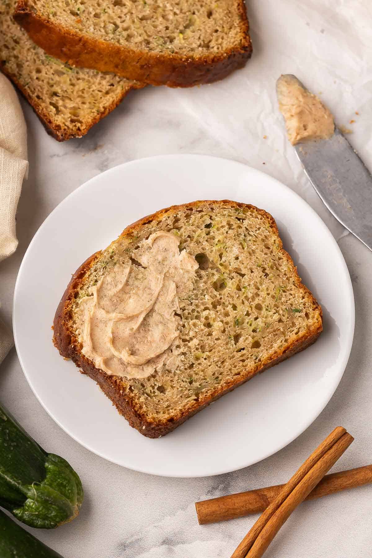 This Perfect Zucchini Bread is a soft, cinnamon-scented one-bowl recipe that comes together in 15 minutes! It’s perfectly moist, lightly sweet, and made with Greek yogurt for a tender crumb. #zucchini #baking #recipes | baking recipes | zucchini recipes | quick bread | healthy baking recipe | a partially buttered slice of zucchini bread on a white plate with cinnamon sticks beside.