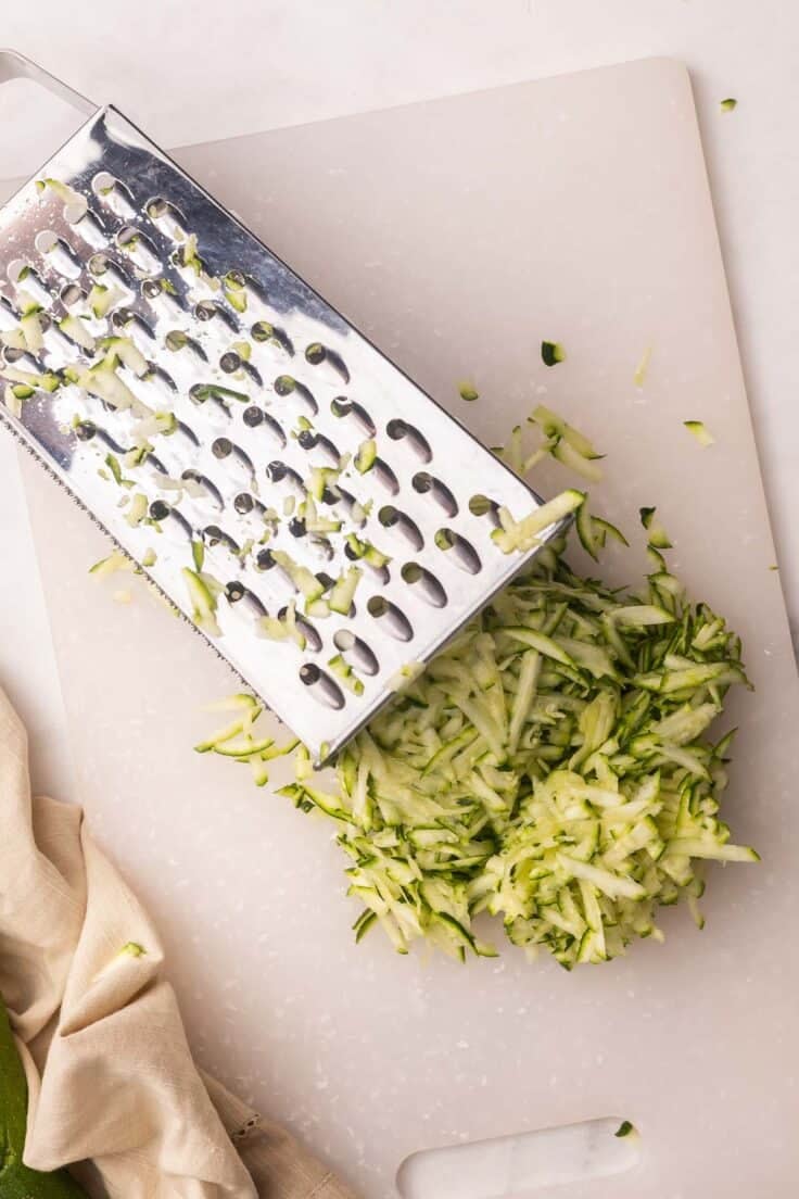 shredded zucchini and shredder on a white cutting board.