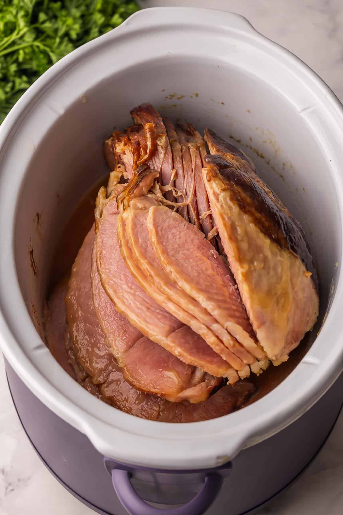 This Crockpot Ham with Pineapple Brown Sugar Glaze is tender, juicy, and incredibly simple. You only need 5 minutes of prep, while the slow cooker takes care of the rest for a flavorful main dish that fits holidays or weeknights! #slowcooker #crockpot #dinner #recipe | easy dinner recipes | dinner ideas | ham recipe | christmas dinner | easter dinner | ham recipe | spiral ham glazed ham in crockpot with parsley beside.