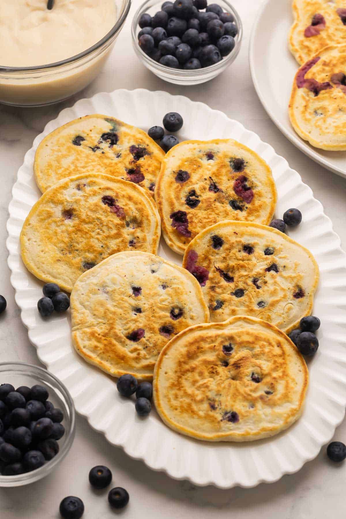 Blueberries and Cream Pancakes are the perfect “treat yourself” stack, fluffy blueberry pancakes covered in warm vanilla custard and extra berries. Ready in 35 minutes and gone even faster! #recipe #pancakes #breakfast | blueberry recipes | pancake recipes | brunch recipes | breakfast recipes | easter brunch | mothers day | sweet breakfast white platter of pancakes with blueberries around it.