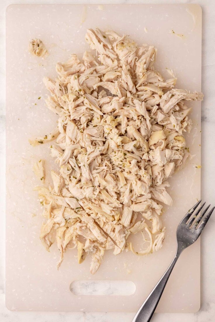 shredded chicken and one fork on a white cutting board.