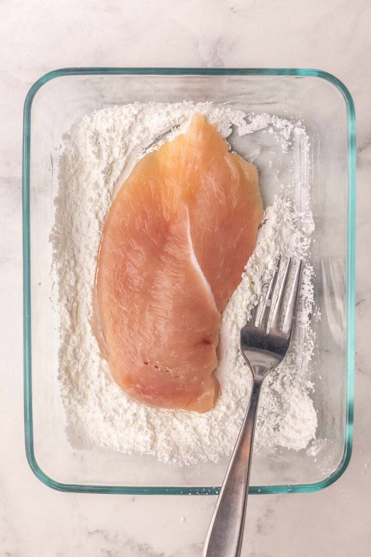 chicken breast in a glass bowl of flour mixture with a fork.