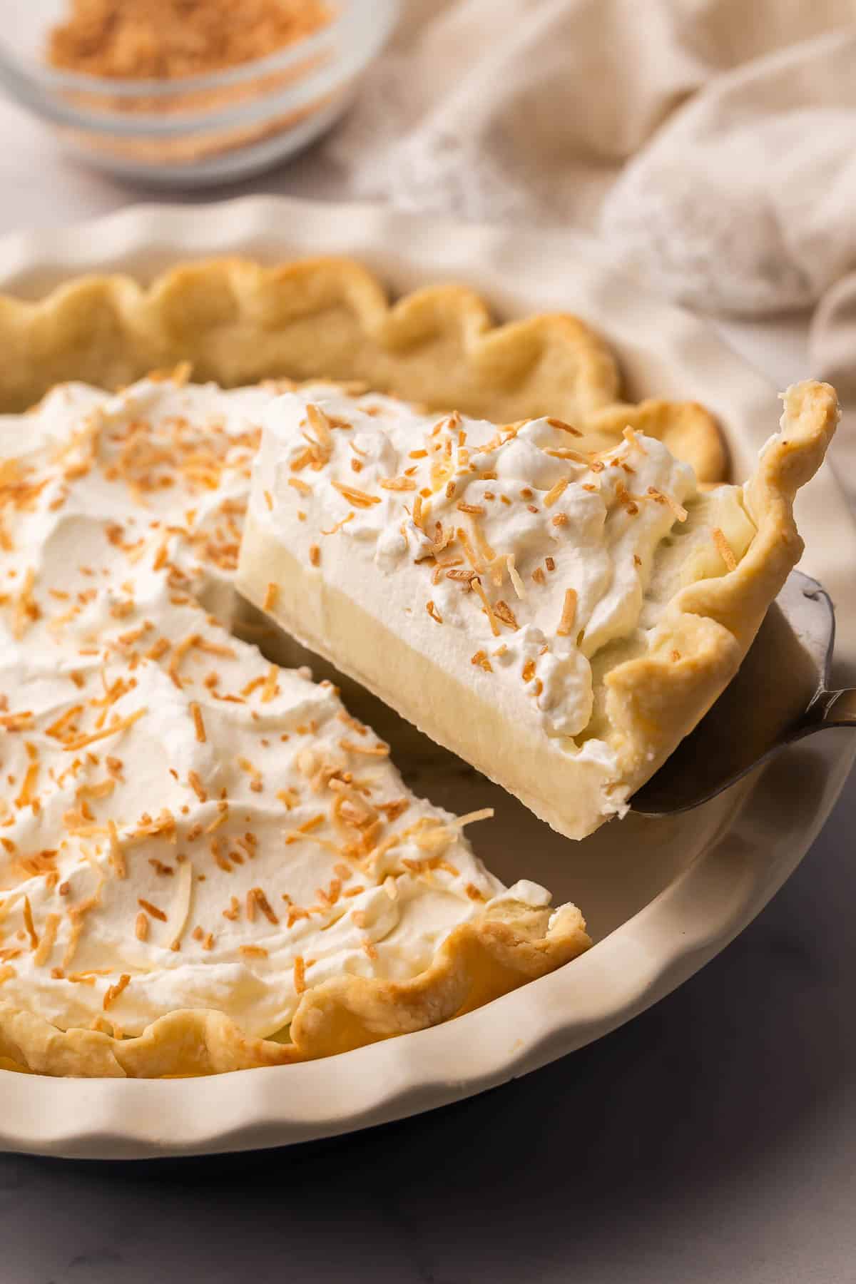 Coconut Cream Pie is a from-scratch dessert with a buttery homemade crust and thick coconut custard filling, finished with whipped cream and toasted coconut. Perfect for spring or Easter dessert! #easter #dessert #recipe | easter recipes | coconut dessert | coconut recipes | coconut pie | easter pie | homemade pie recipe | easter dessert a piece of coconut cream pie being lifted out of pie dish.