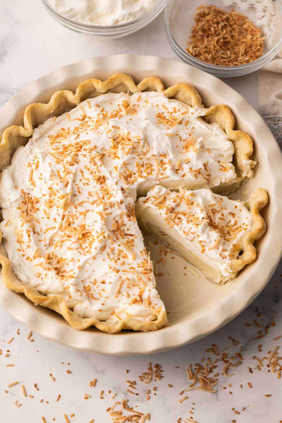 Coconut Cream Pie is a from-scratch dessert with a buttery homemade crust and thick coconut custard filling, finished with whipped cream and toasted coconut. Perfect for spring or Easter dessert! #easter #dessert #recipe | easter recipes | coconut dessert | coconut recipes | coconut pie | easter pie | homemade pie recipe | easter dessert a slice of coconut cream pie in pan with toasted coconut sprinkled around.