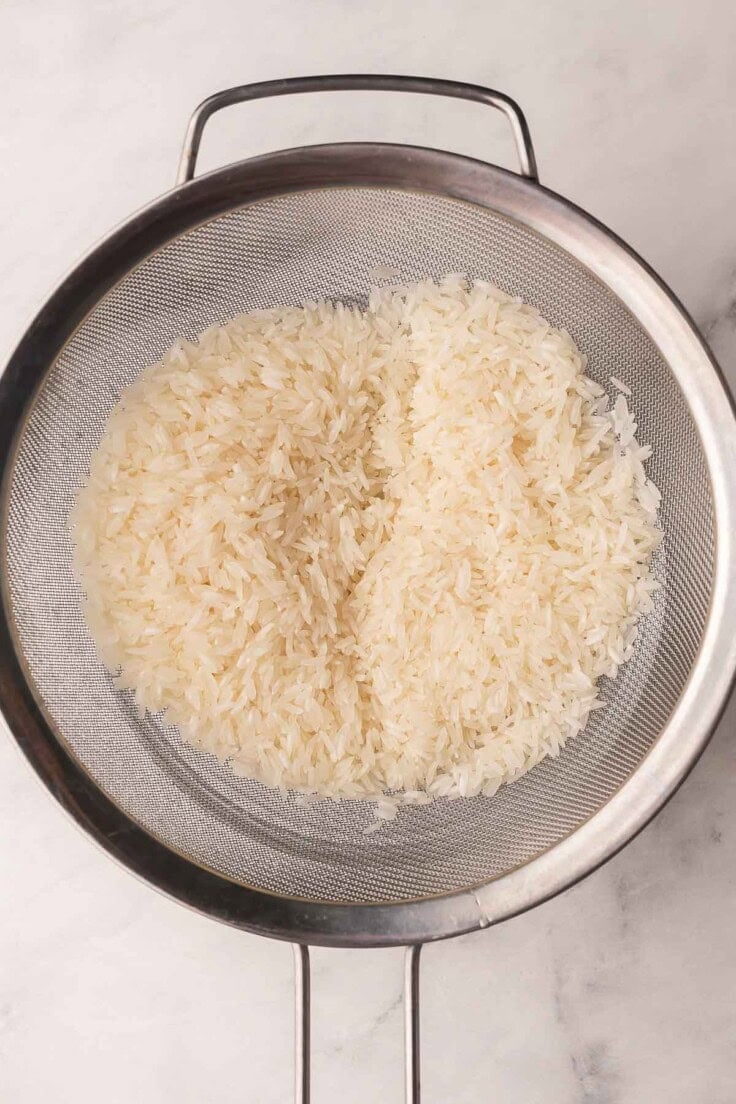 long grain white rice being rinsed in a strainer.