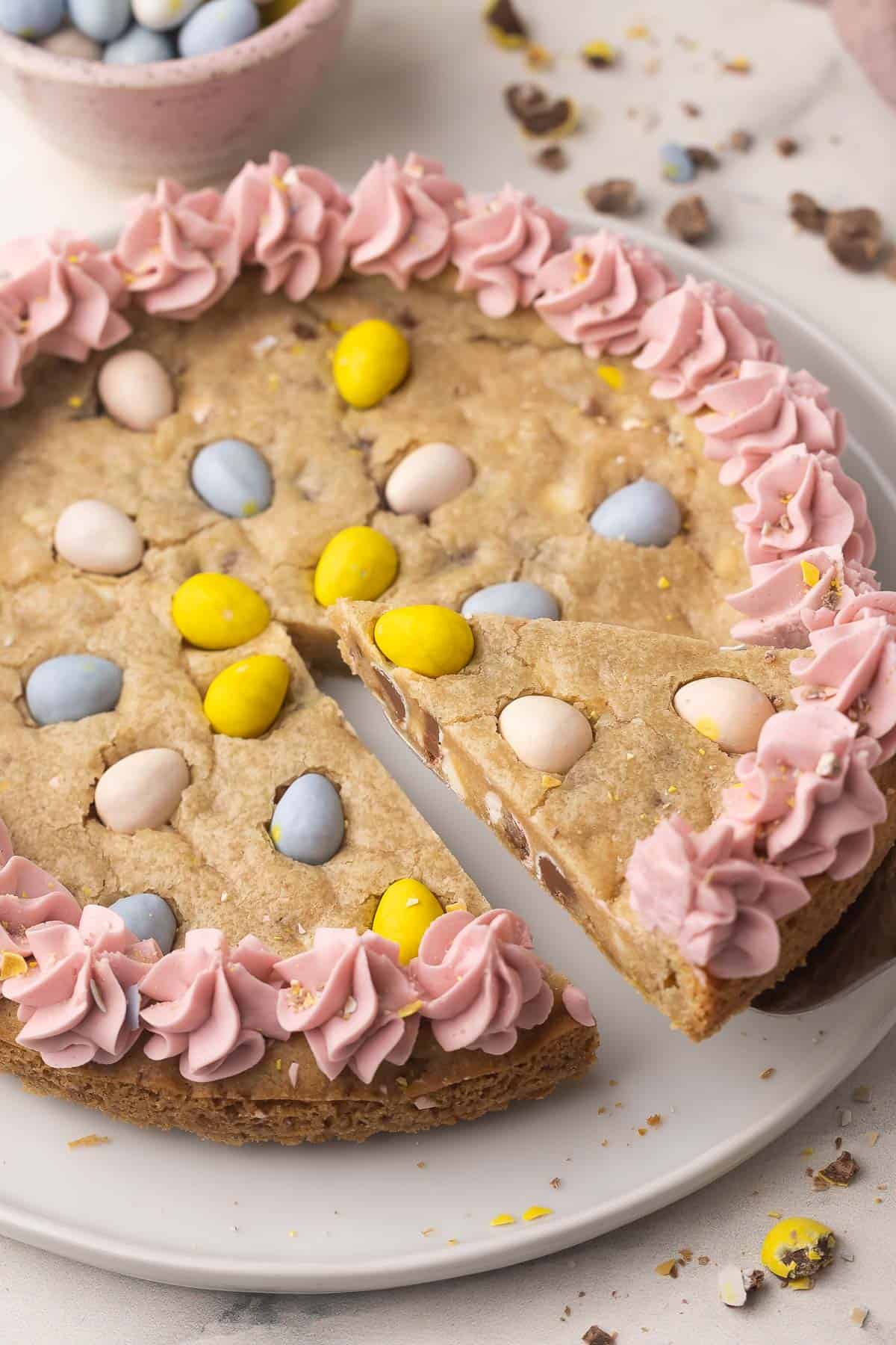Make this Easter Mini Egg Cookie Cake in 30 minutes, packed with Mini Eggs and white chocolate, finished with pretty pastel buttercream swirls! An easy Easter dessert that disappears quickly. #easter #dessert #cookie | cookie recipes | cookie cake | skillet cookie | easter cookies | easter cake | easter recipes | mini egg recipes | easter eggs | chocolate chip cookie a slice of mini egg cookie cake being lifted up.