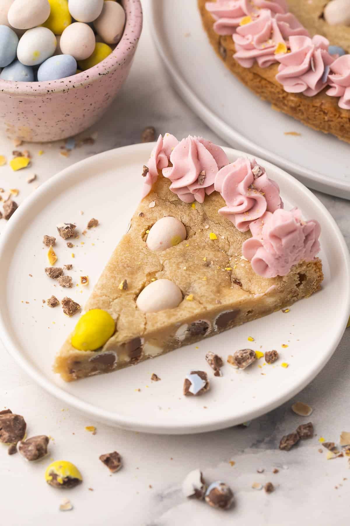 Make this Easter Mini Egg Cookie Cake in 30 minutes, packed with Mini Eggs and white chocolate, finished with pretty pastel buttercream swirls! An easy Easter dessert that disappears quickly. #easter #dessert #cookie | cookie recipes | cookie cake | skillet cookie | easter cookies | easter cake | easter recipes | mini egg recipes | easter eggs | chocolate chip cookie a slice of mini egg cookie cake on a white plate with mini eggs beside.