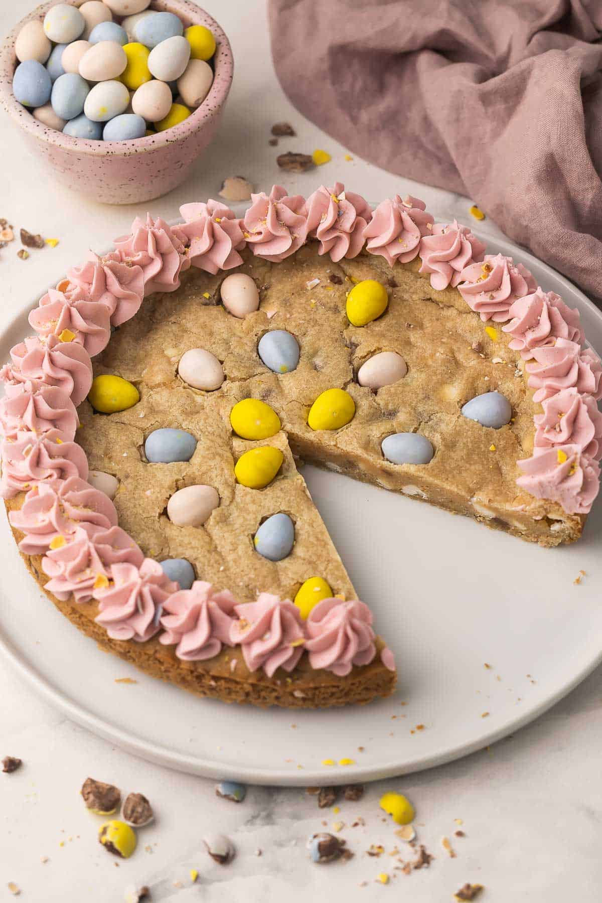 Make this Easter Mini Egg Cookie Cake in 30 minutes, packed with Mini Eggs and white chocolate, finished with pretty pastel buttercream swirls! An easy Easter dessert that disappears quickly. #easter #dessert #cookie | cookie recipes | cookie cake | skillet cookie | easter cookies | easter cake | easter recipes | mini egg recipes | easter eggs | chocolate chip cookie mini egg cookie cake with a slice missing and mini eggs beside.