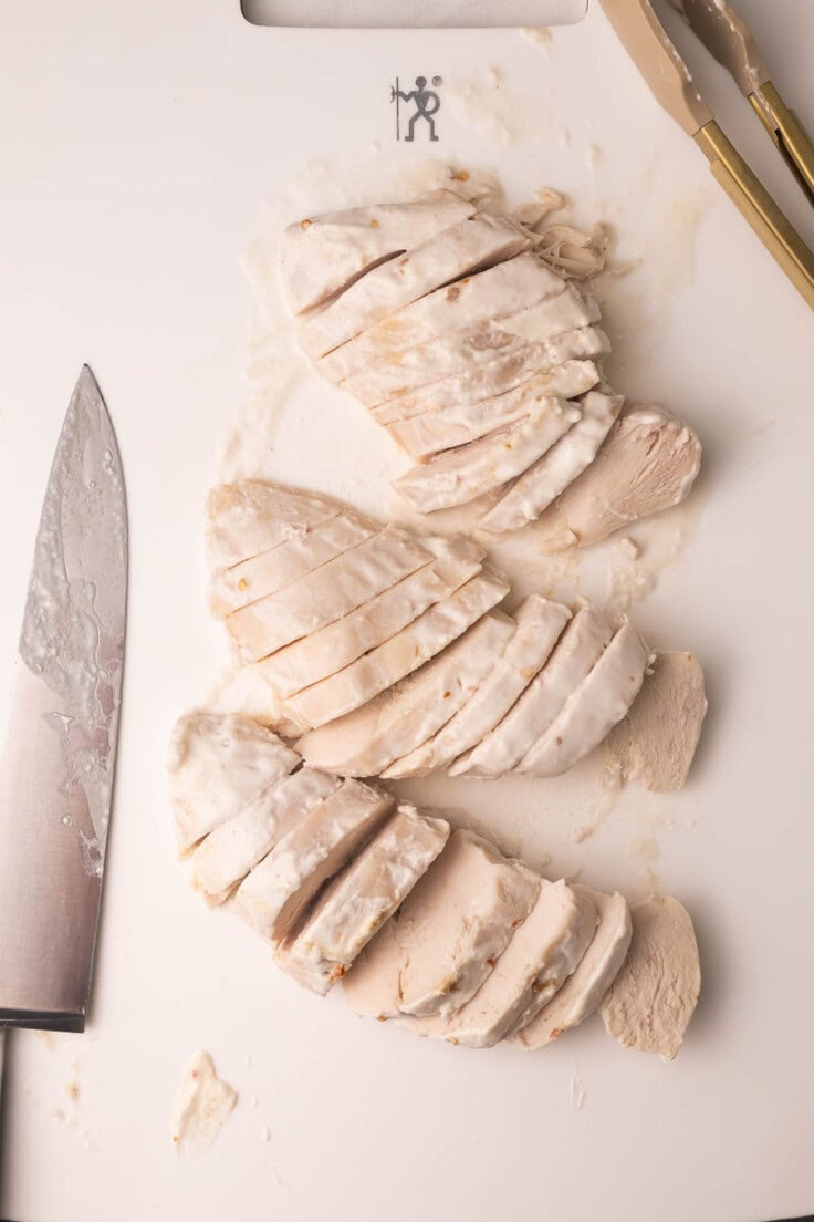 three cooked chicken breast sliced on a white cutting board.