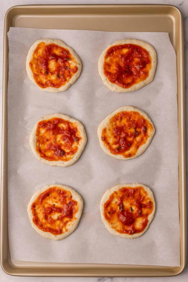 tomato sauce spread on pizza bun dough on a parchment lined pan.