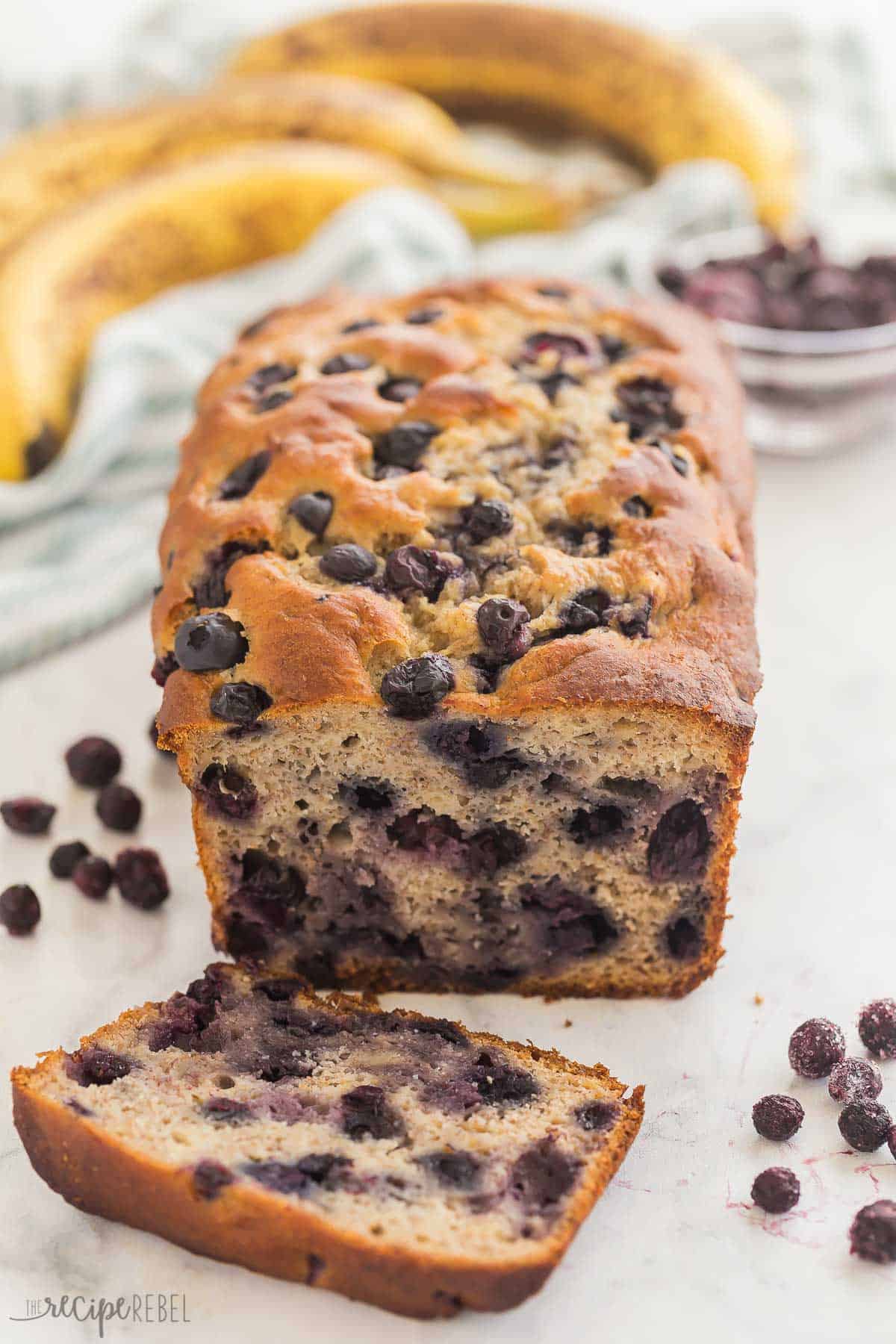 This Blueberry Banana Bread is ultra moist and tender, loaded with bananas and blueberries! Use fresh or frozen blueberries and fresh or frozen bananas -- it's perfect for breakfast on the go. #banana #blueberry #baking #recipe | banana bread recipes | blueberry recipes | banana recipes | baking recipes | breakfast ideas | on the go breakfast | kids lunch one slice cut off of a loaf of blueberry banana bread with bananas in the background.