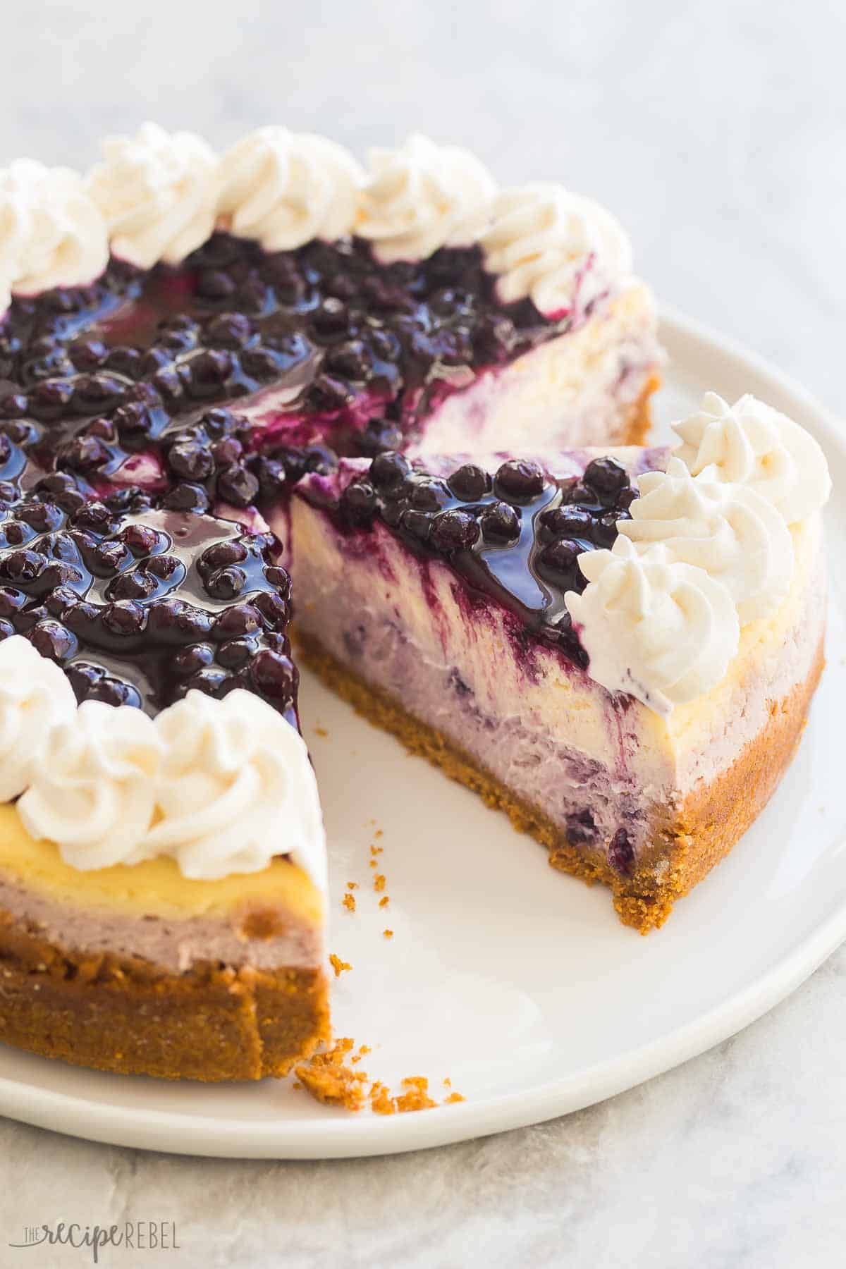 This Blueberry Cheesecake is ultra creamy and swirled with fresh blueberry sauce. It's made with no water bath and no fuss and is the perfect summer dessert! #blueberry #cheesecake #dessert #recipe | dessert recipes | cheesecake recipes | blueberry desserts | easy cheesecake | easy desserts | summer recipes one piece cut from the whole blueberry cheesecake with whipped cream.