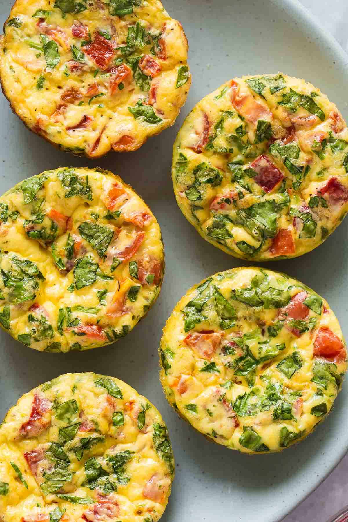 These Mini Frittata Muffins are an easy breakfast recipe made with eggs, cream cheese, spinach, tomatoes and roasted red peppers. They're high in protein and meal prep, make ahead and freezer friendly! #frittata #eggs #recipe #breakfast | breakfast recipes | breakfast ideas | egg muffins | egg bites | protein recipes | protein breakfast | healthy recipes overhead image of frittata muffins on a blue plate.