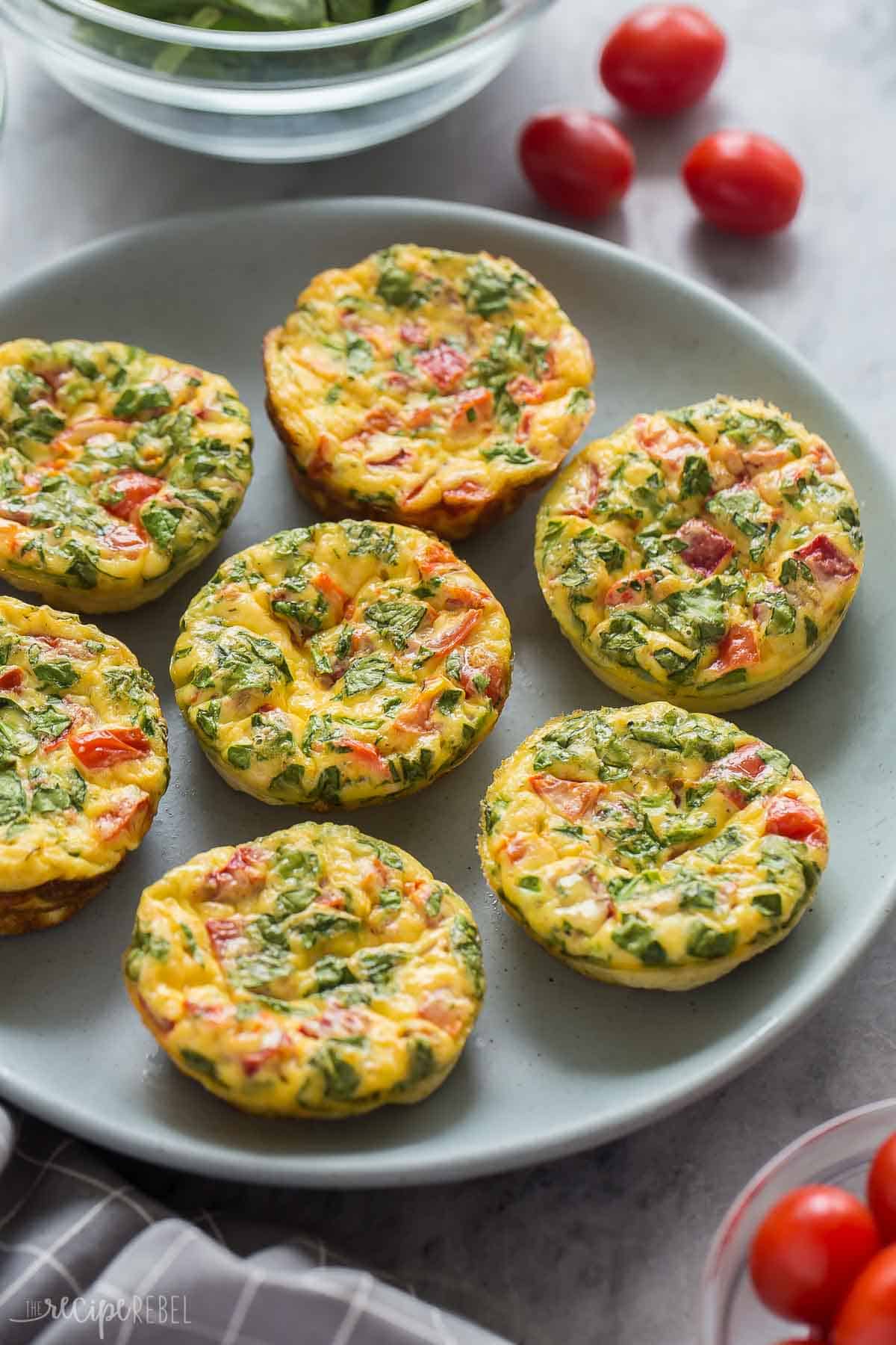 These Mini Frittata Muffins are an easy breakfast recipe made with eggs, cream cheese, spinach, tomatoes and roasted red peppers. They're high in protein and meal prep, make ahead and freezer friendly! #frittata #eggs #recipe #breakfast | breakfast recipes | breakfast ideas | egg muffins | egg bites | protein recipes | protein breakfast | healthy recipes spinach and tomato egg muffins on a blue plate with tomatoes behind.