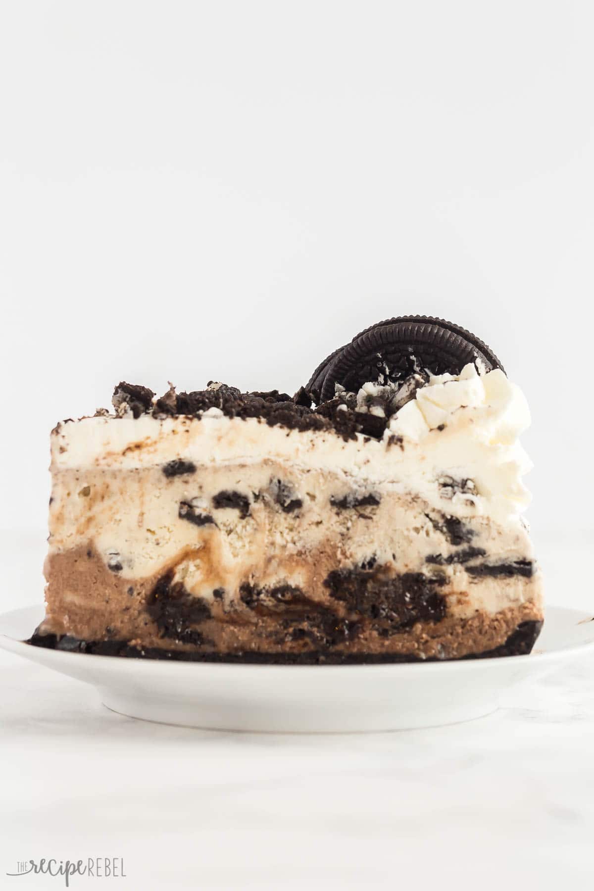 This Oreo Ice Cream Cake is made with an Oreo crust, chocolate ice cream, vanilla ice cream, a rich fudge layer and topped with whipped cream -- for Oreo lovers only! #oreo #icecream #dessert #recipe | oreo desserts | ice cream desserts | ice cream recipes | oreo recipes | summer dessert | easy dessert slice of oreo ice cream cake on a white plate with whipped cream on top.