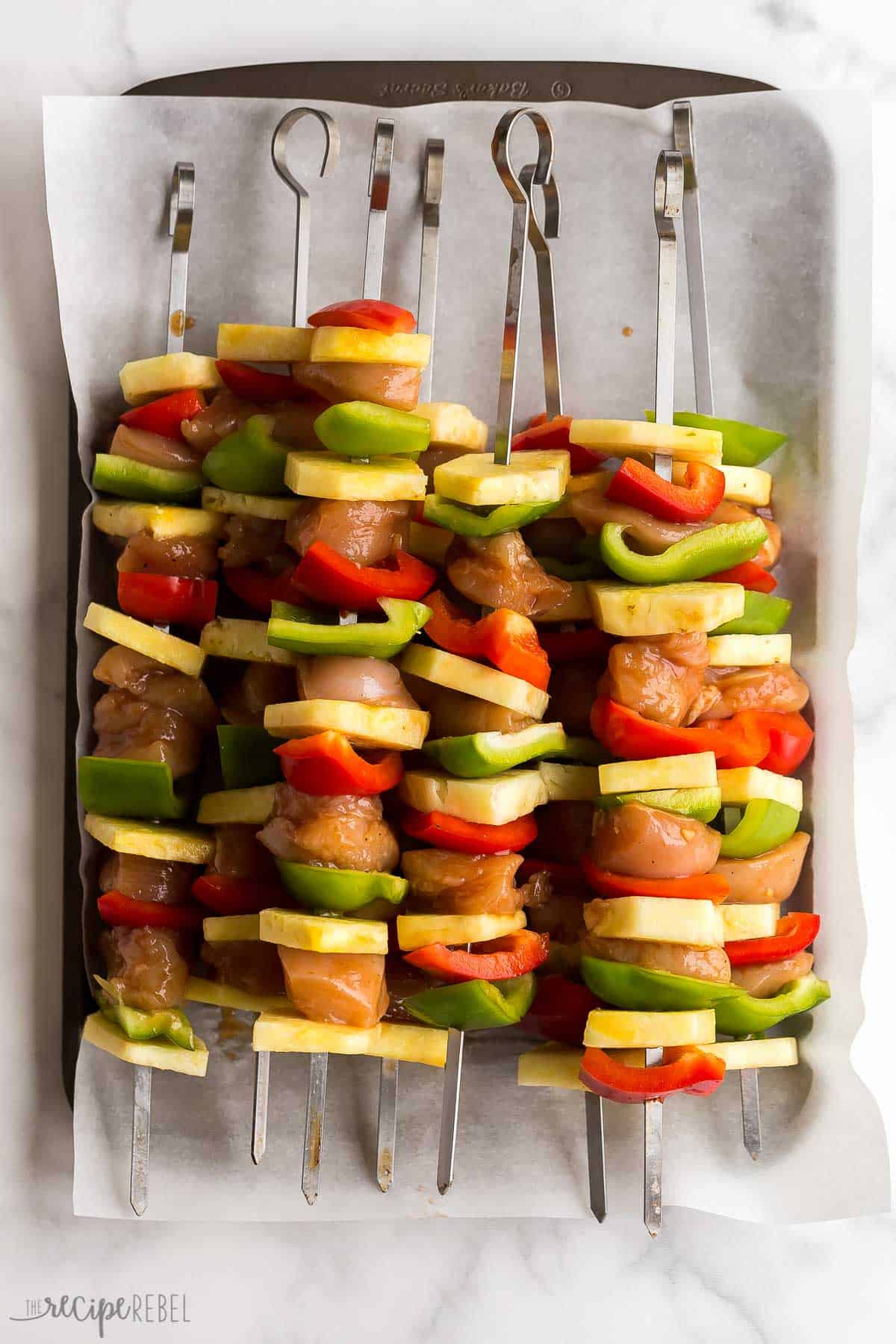 These Hawaiian Chicken Kabobs are sweet and savory, loaded with tender chicken breast, pineapple chunks, bell peppers and an easy pineapple BBQ marinade! The perfect prep-ahead summer meal. #chicken #chickenbreast #kabob #grilling #dinner | easy dinner ideas | dinner recipes | chicken recipes | grilled chicken | summer grilling | summer dinner uncooked hawaiian chicken kabobs on sheet pan with pineapple and peppers.