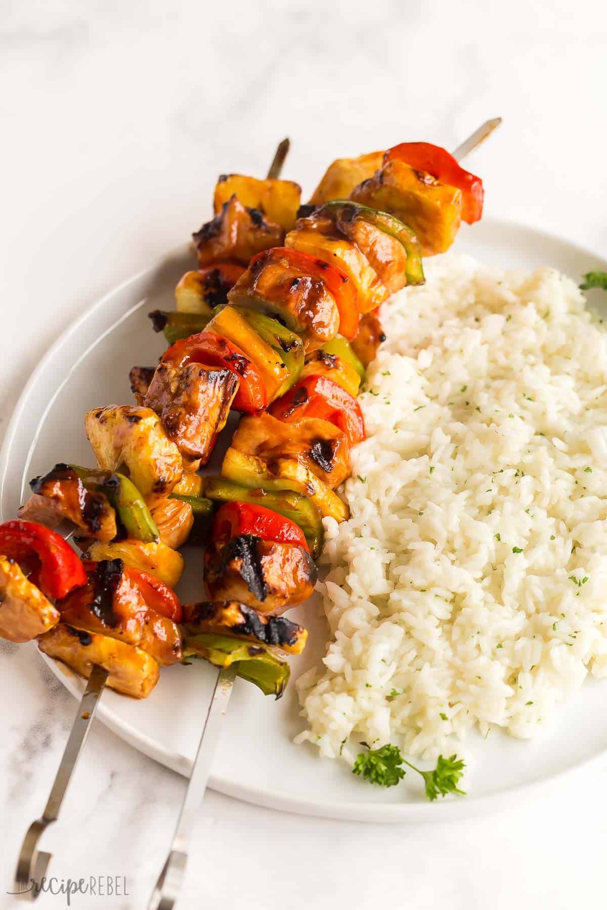 These Hawaiian Chicken Kabobs are sweet and savory, loaded with tender chicken breast, pineapple chunks, bell peppers and an easy pineapple BBQ marinade! The perfect prep-ahead summer meal. #chicken #chickenbreast #kabob #grilling #dinner | easy dinner ideas | dinner recipes | chicken recipes | grilled chicken | summer grilling | summer dinner three metal skewers with hawaiian chicken and peppers on a plate of white rice.