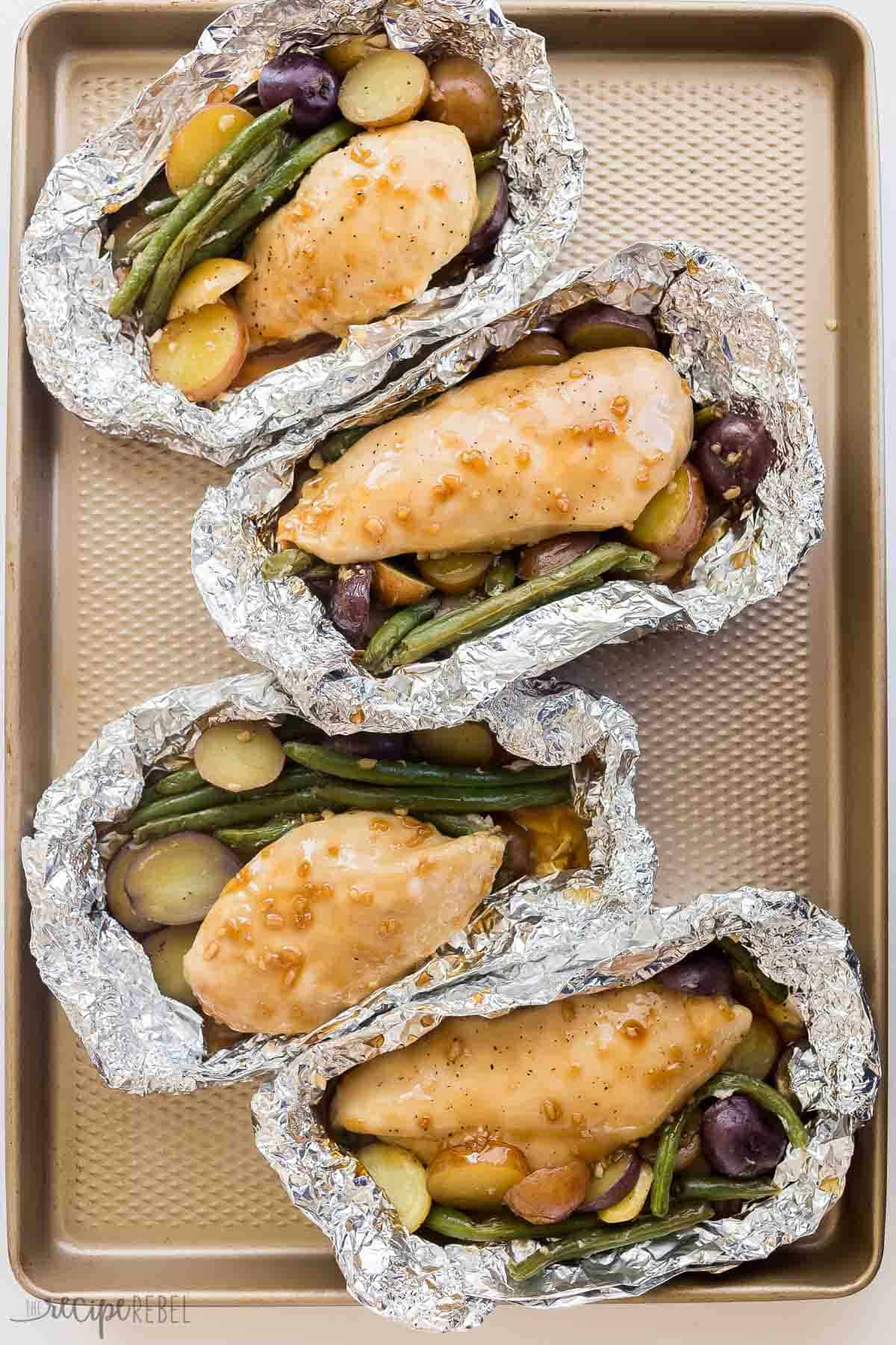 These Honey Garlic Chicken Foil Packets are made with juicy grilled chicken breast, potatoes and green beans and covered in the simplest homemade honey garlic sauce -- an easy camping meal perfect for summer dinners! overhead image of honey garlic chicken foil packs on a baking sheet.