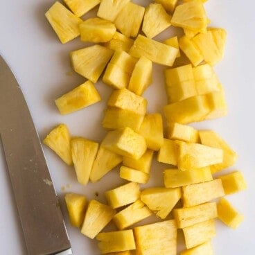 pineapple cut into small pieces on a white cutting board with a knife.